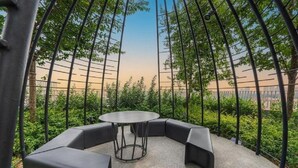 Un espace extérieur circulaire aménagé avec des sièges, une table centrale et des bancs disposés autour, le tout entouré d’une clôture en métal et de verdure.