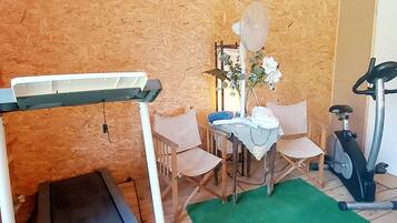 A room with a treadmill, a dining table with chairs, a clock on the wall, and a green rug.