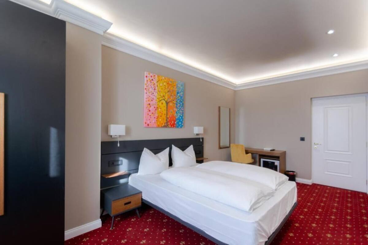 Habitación de hotel con una cama grande, mesitas de noche, un escritorio, una silla, un cuadro en la pared y una puerta.