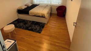 A small bedroom with a bed, a window with blinds, a red chair, a small stool, and a heater.