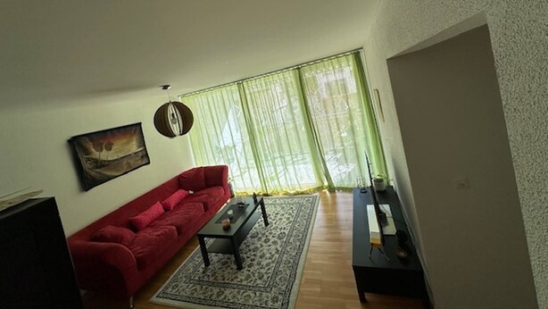 A living room with a red sofa, a coffee table, a painting on the wall, and a large window with sheer curtains.