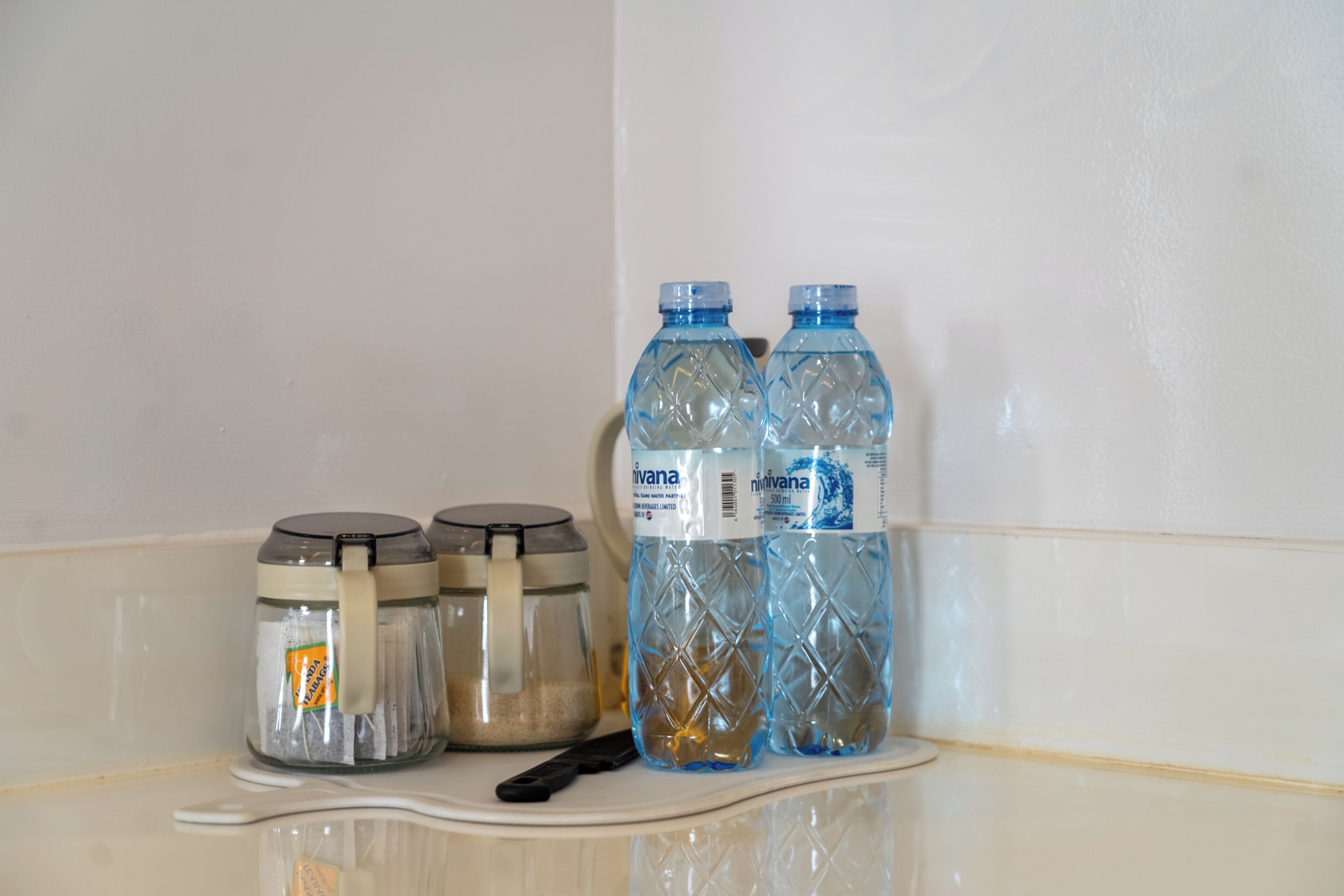 Une salle de bain avec un mur recouvert de carreaux blancs, deux bocaux en verre avec des couvercles et deux bouteilles d’eau sur une étagère blanche.