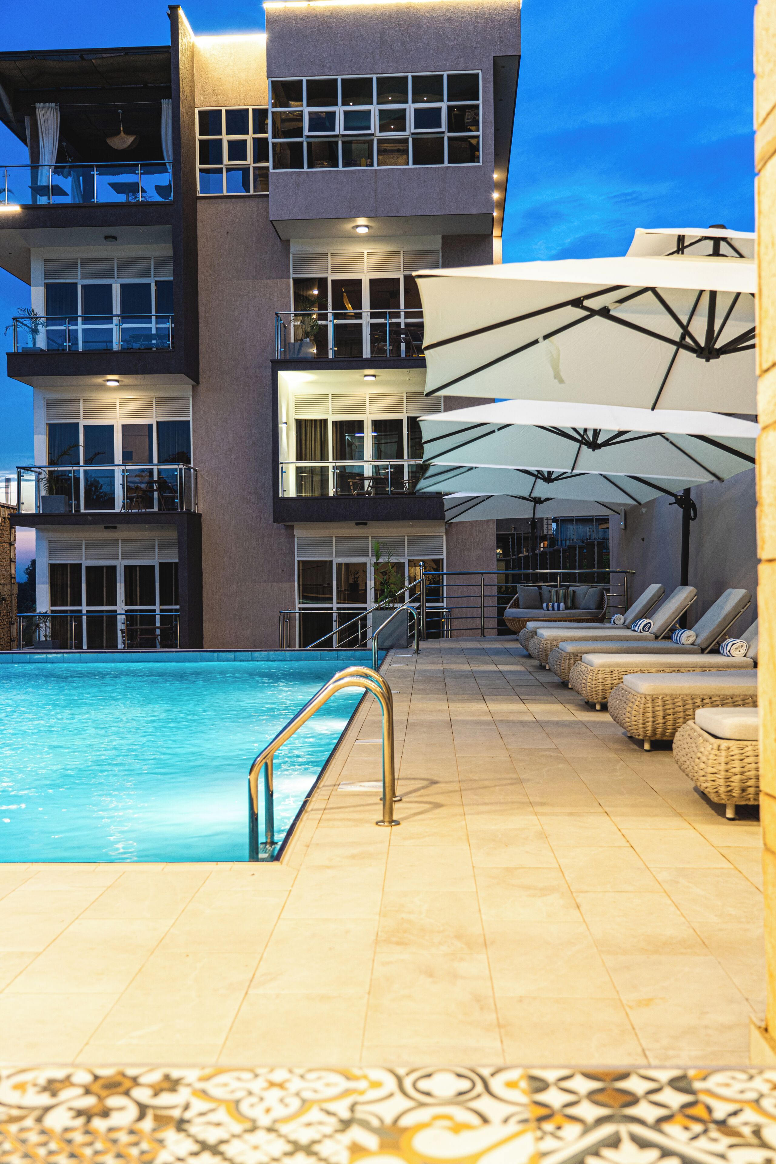 A swimming pool with sun loungers and umbrellas, a multi-story building with balconies, and a tiled floor.