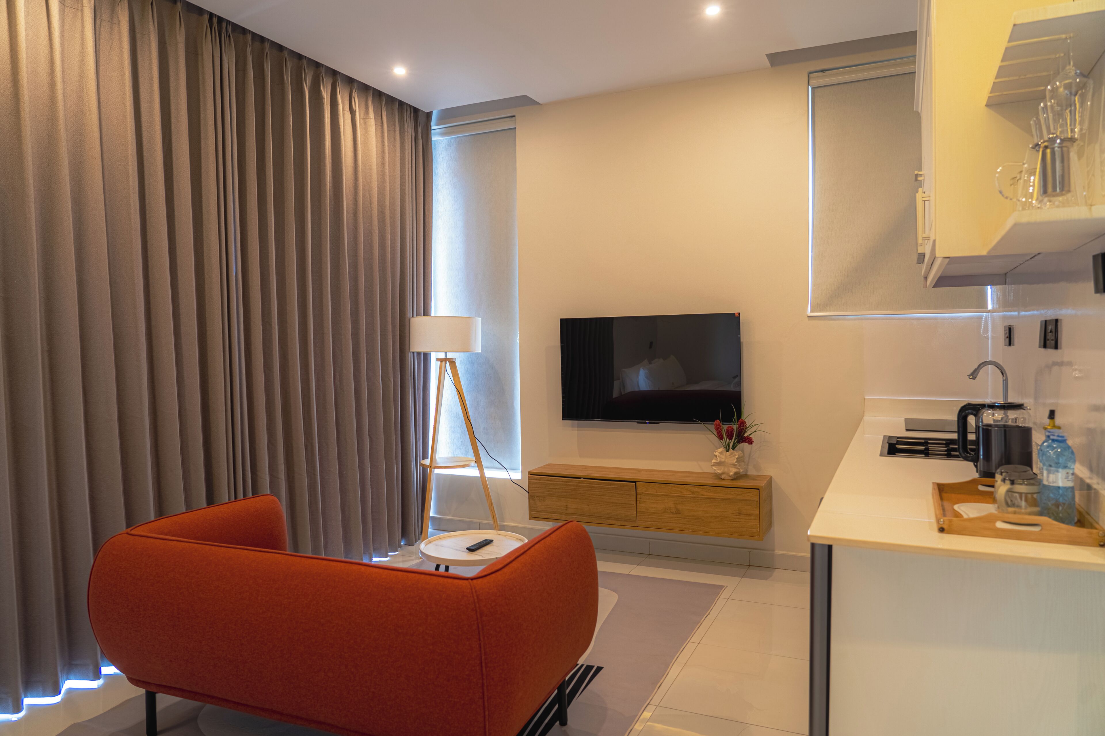 A modern hotel room with a sofa, a TV, a bedside table, a lamp, a kitchen area, and a large window with curtains.