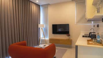 A modern hotel room with a sofa, a TV, a bedside table, a lamp, a kitchen area, and a large window with curtains.