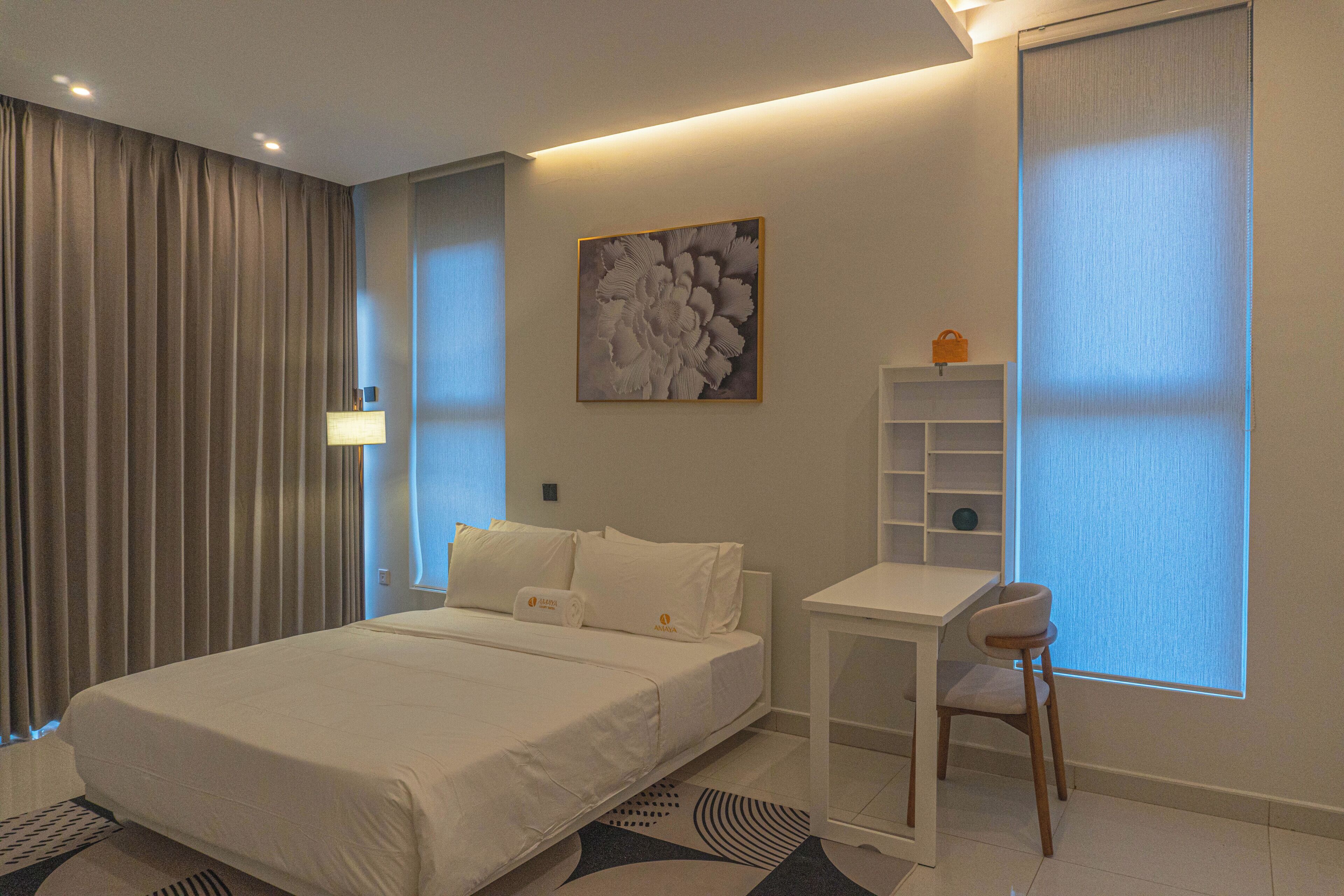A modern hotel room with a bed, a desk, a chair, a large window with blinds, a wall-mounted shelf, and a painting.