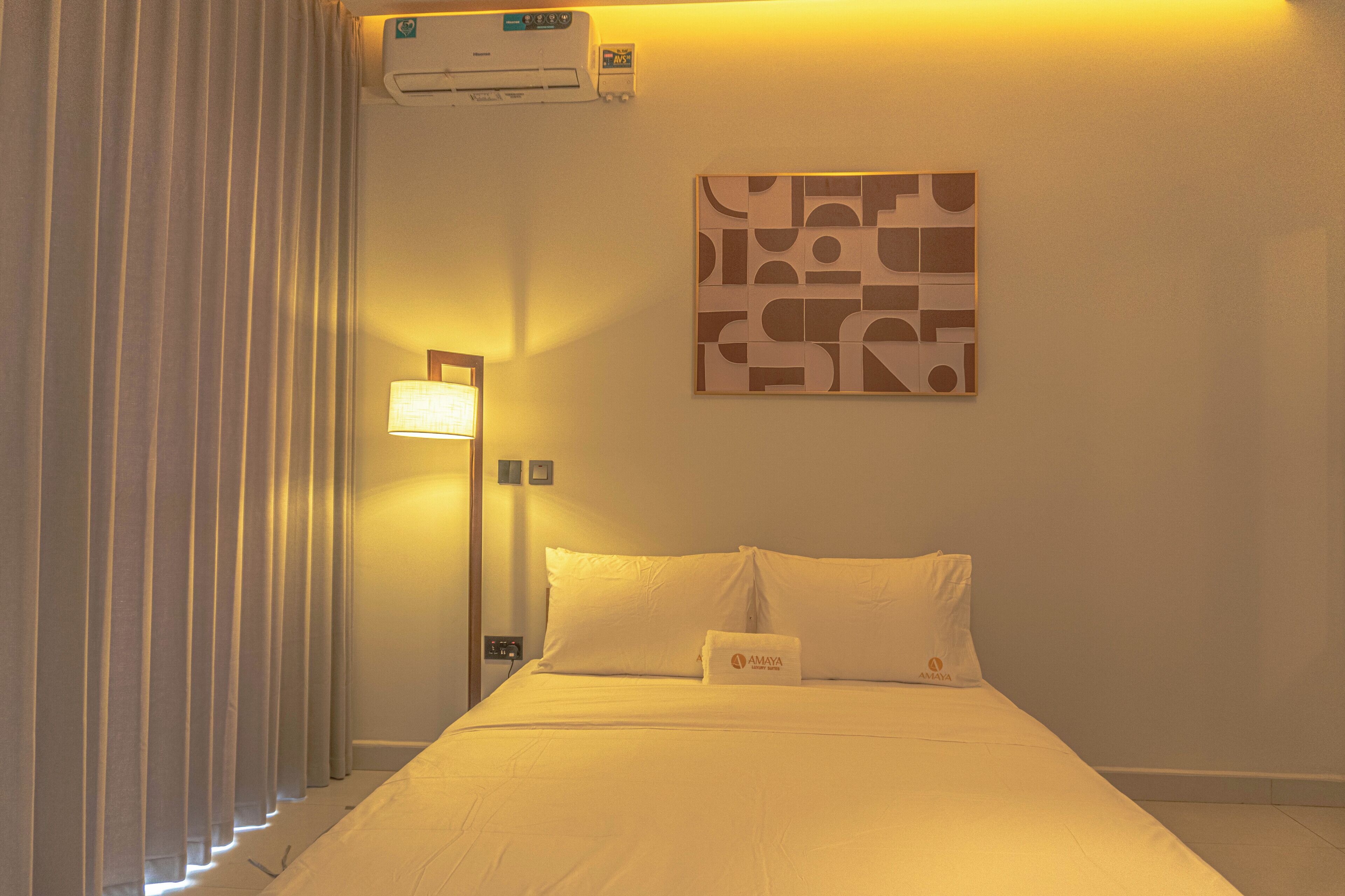 A hotel room with a bed, a wall-mounted air conditioner, a bedside lamp, and a decorative wall piece.