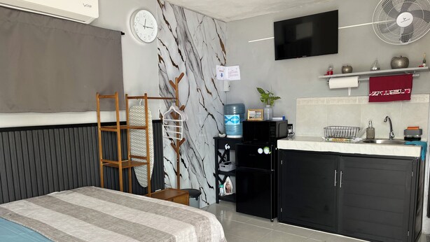 Una habitación pequeña con cama, kitchenette, televisor, dispensador de agua y reloj.