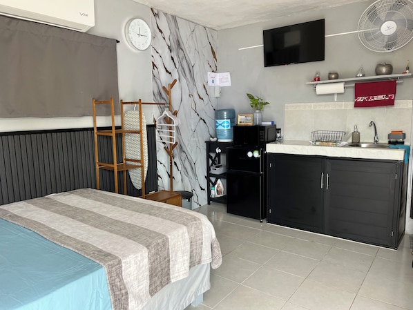 A compact room with a bed, a small kitchenette, a TV, a water dispenser, and a clock.