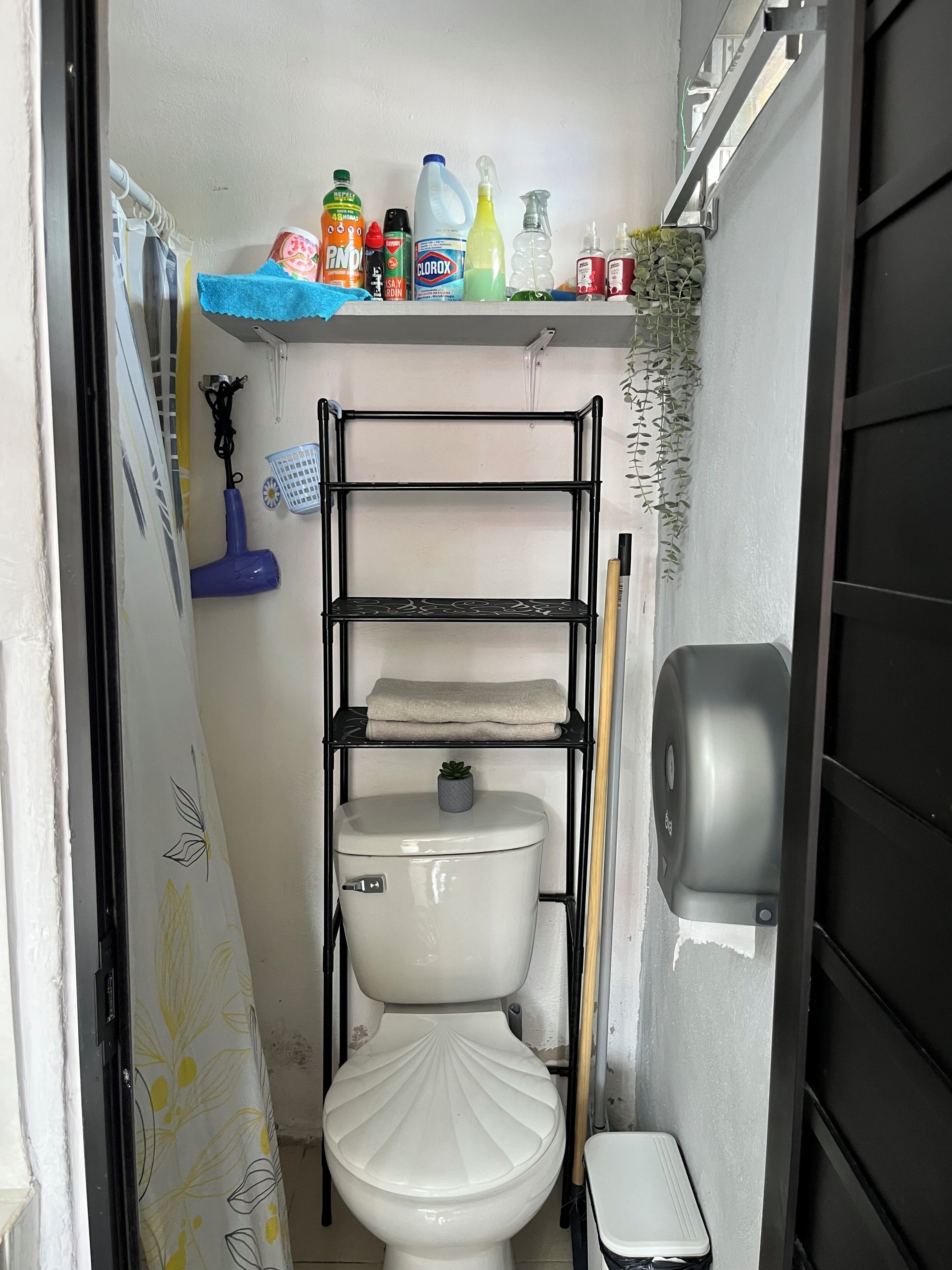 A small bathroom with a toilet, a shelf with cleaning supplies, a hanging basket, and a hand dryer.