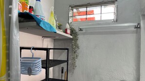 Um quarto pequeno e estreito com uma janela, uma prateleira com produtos de limpeza, uma cesta azul e uma toalha sobre uma estante.
