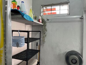 Une petite pièce étroite avec une fenêtre, une étagère contenant des produits de nettoyage, un panier bleu et une serviette posée sur une autre étagère.