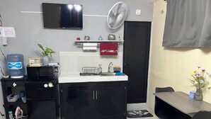Cocina compacta con microondas, dispensador de agua y un refrigerador pequeño. Un televisor está montado en la pared y un ventilador encima del lavamanos.