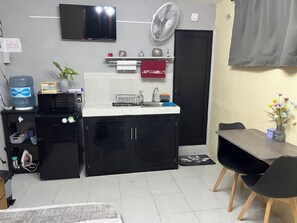 A compact kitchen with a microwave, water dispenser, and a small refrigerator. A TV is mounted on the wall, and a fan is placed above the sink.