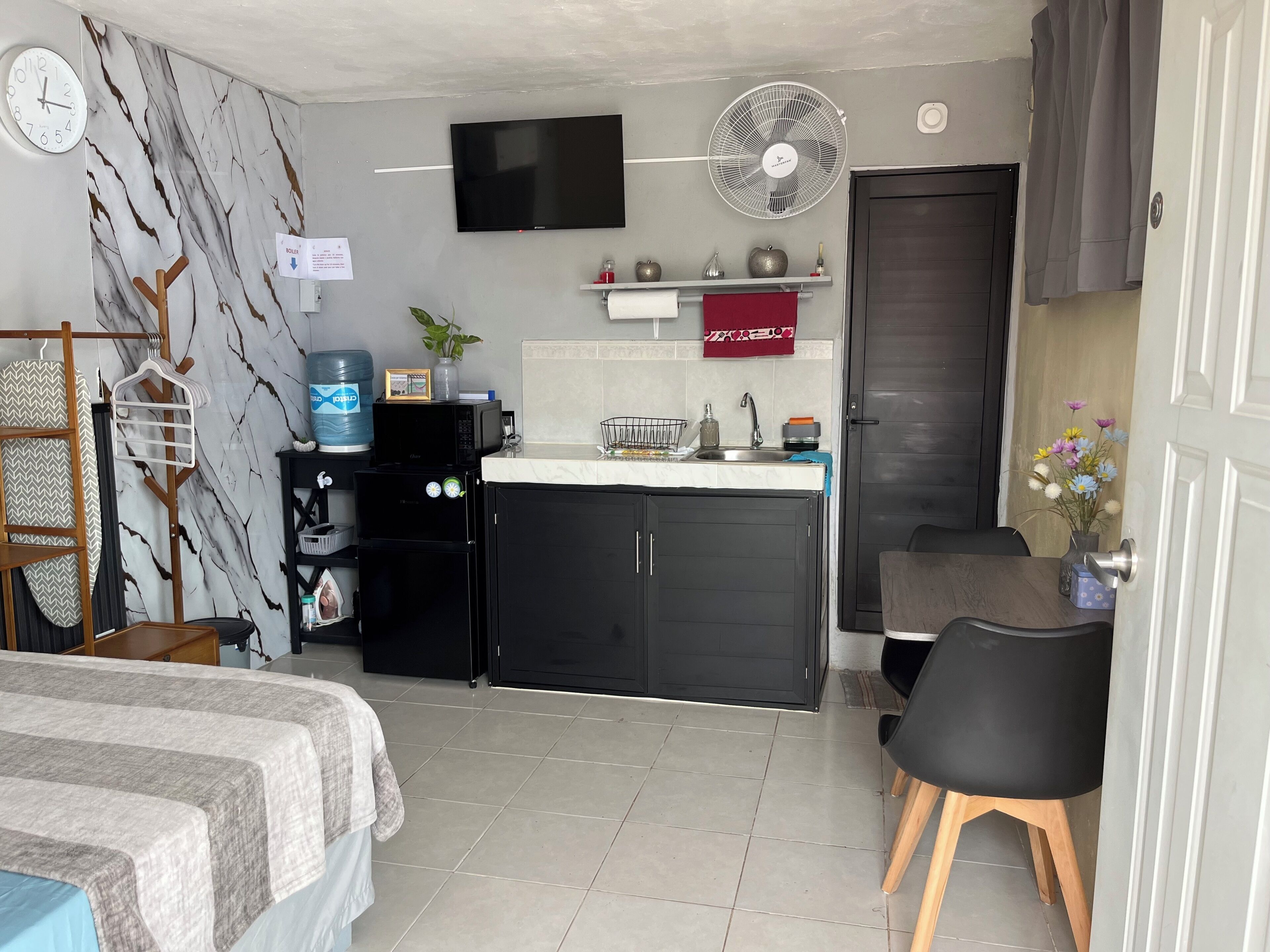 Habitación de hotel compacta con cama, kitchenette, televisor, ventilador y puerta de baño.