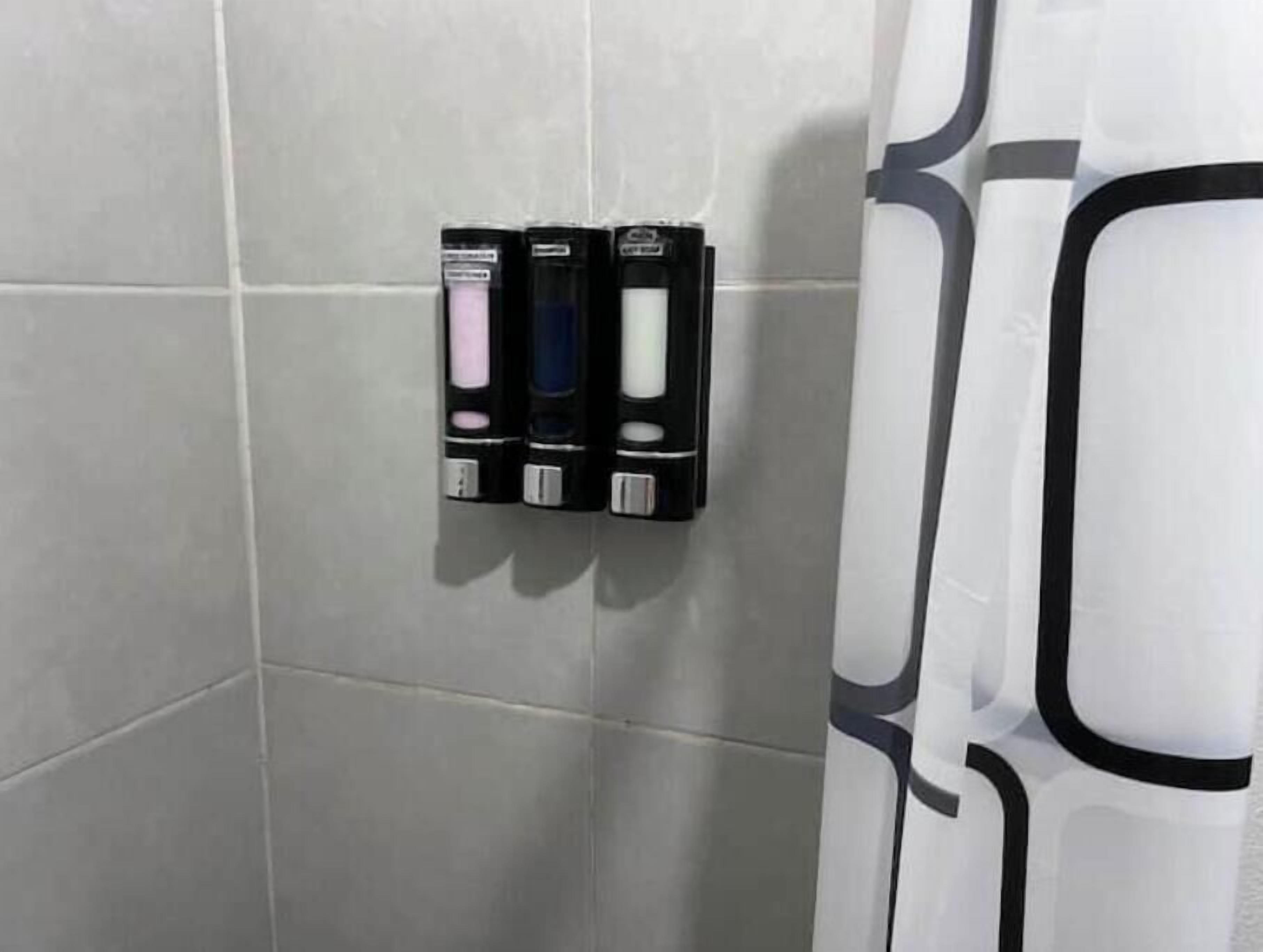 Un baño con azulejos grises, un dispensador de jabón negro y una cortina de ducha blanca con un diseño negro.