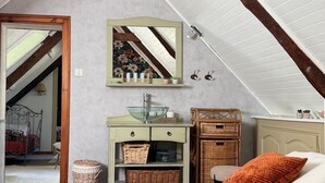 Une chambre avec un plafond en pente, un lit recouvert d’une couvre-lit à motifs, une commode avec des paniers, un miroir et un fauteuil en osier.