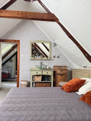 Ein Schlafzimmer mit geneigter Decke, einem Bett mit gemustertem Überwurf, einer Kommode mit Körben, einem Spiegel und einem Korbstuhl.