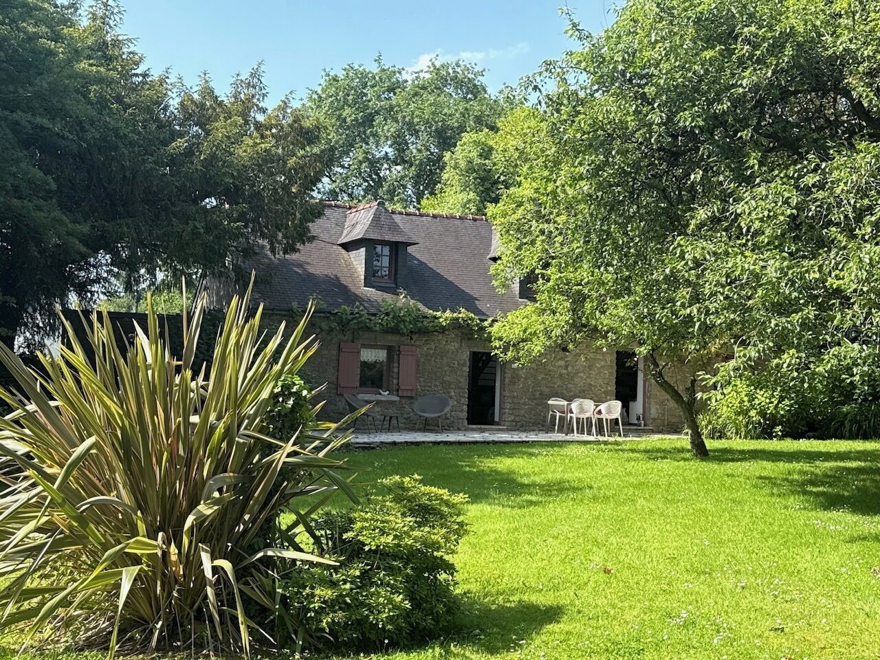 A stone house with a small garden, a patio with chairs, and a tree in front.