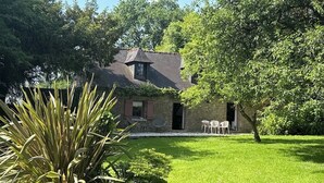 Une maison en pierre avec un petit jardin, une terrasse avec des chaises et un arbre devant.