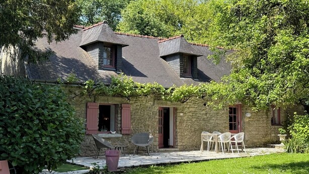 Une maison en pierre avec un toit en pente, deux fenêtres avec volets, un petit porche avec des chaises en osier et une cour gazonnée.
