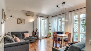 A spacious living area with a grey sofa, a dining table with chairs, and large windows with curtains.