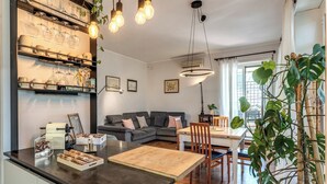 A modern kitchen with a central island, a dining area with a table and chairs, a gray sofa, and a large window with natural light.