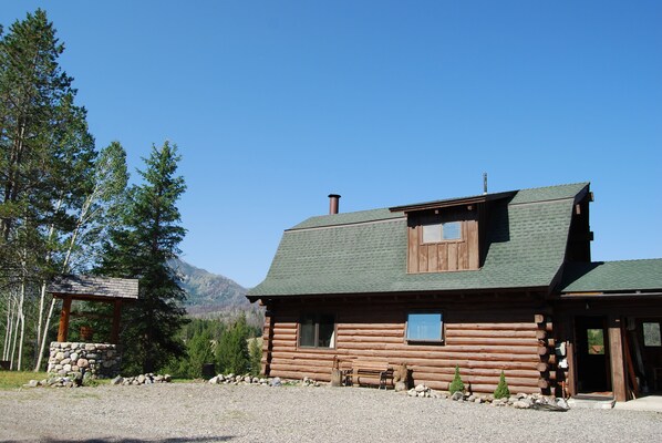 A log cabin with a stone foundation, a wooden porch, and a mountain backdrop.