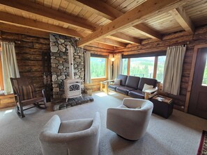 A log cabin interior with a stone fireplace, wooden ceiling beams, a sofa, and a view of the outdoors.