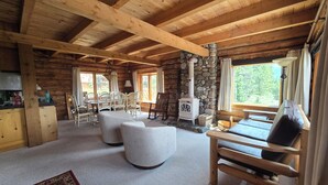 A log cabin interior with a stone fireplace, wooden furniture, and a view of the outdoors.
