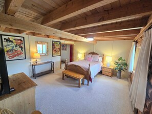 A cozy bedroom with a wooden ceiling, a bed with pink and white bedding, a nightstand with a lamp, and a poster on the wall.