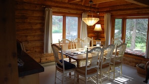 A wooden cabin interior with a dining table surrounded by chairs, a chandelier, and large windows with curtains.