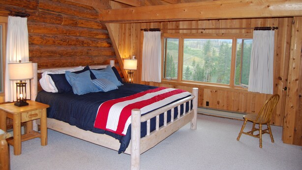A log cabin bedroom with a bed, two bedside lamps, a chair, and a window with a view of trees.