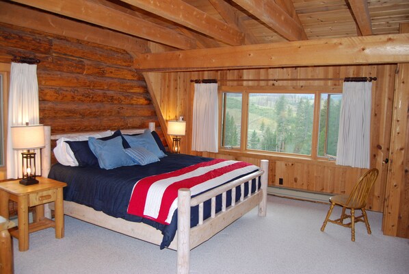 A log cabin bedroom with a bed, two bedside lamps, a chair, and a window with a view of trees.