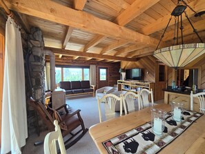 A cozy cabin interior with a stone fireplace, wooden ceiling beams, a dining table with chairs, a sofa, and a television. - Cabin by the Creek in Stanley Basin (Sawtooth City)