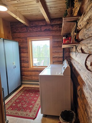 A laundry room with a washing machine, a blue cabinet, a red patterned rug, and a window with a view of trees and buildings. - Cabin by the Creek in Stanley Basin (Sawtooth City)