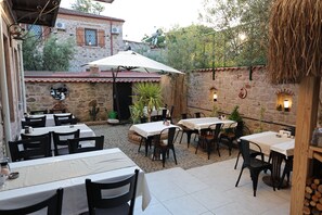 An outdoor patio area with tables and chairs, a thatched roof, stone walls, and a view of a building with a red roof. - Adena Pansiyon (Küçükköy)