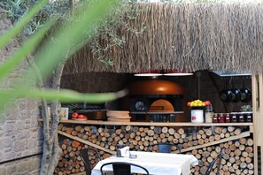 Cocina exterior con pared de ladrillo, techo de paja y mesa con sillas.