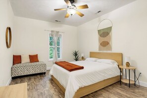 Une chambre à coucher comprenant un lit, une table de chevet, une commode, un ventilateur de plafond, une fenêtre avec des rideaux et une œuvre d’art abstraite accrochée au mur.