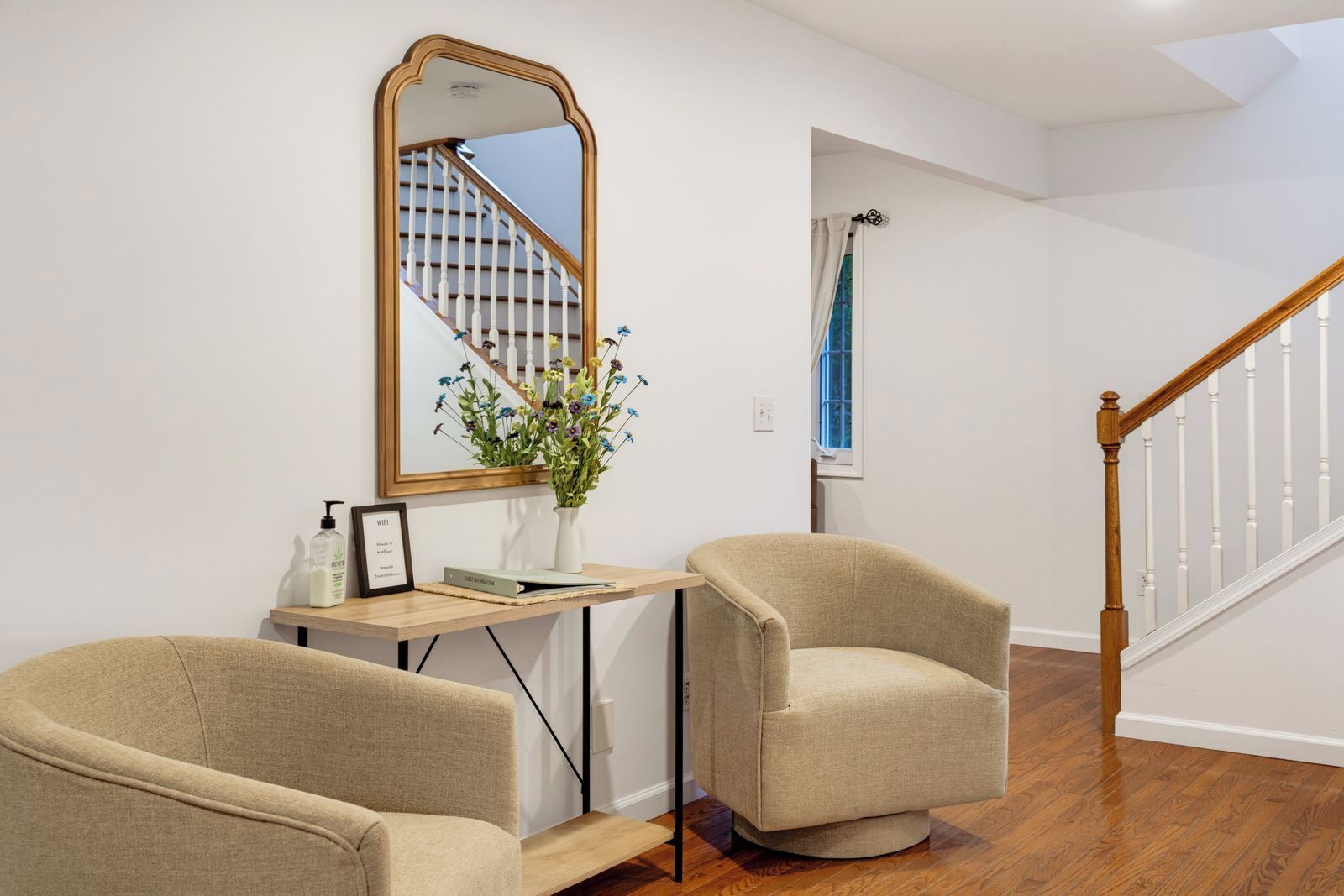 Een kamer met een houten trap, twee fauteuils, een bureau met spiegel, een vaas met bloemen en een lijst met afbeelding.