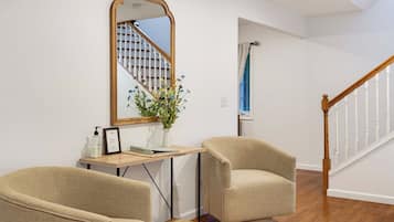 Een kamer met een houten trap, twee fauteuils, een bureau met spiegel, een vaas met bloemen en een lijst met afbeelding.