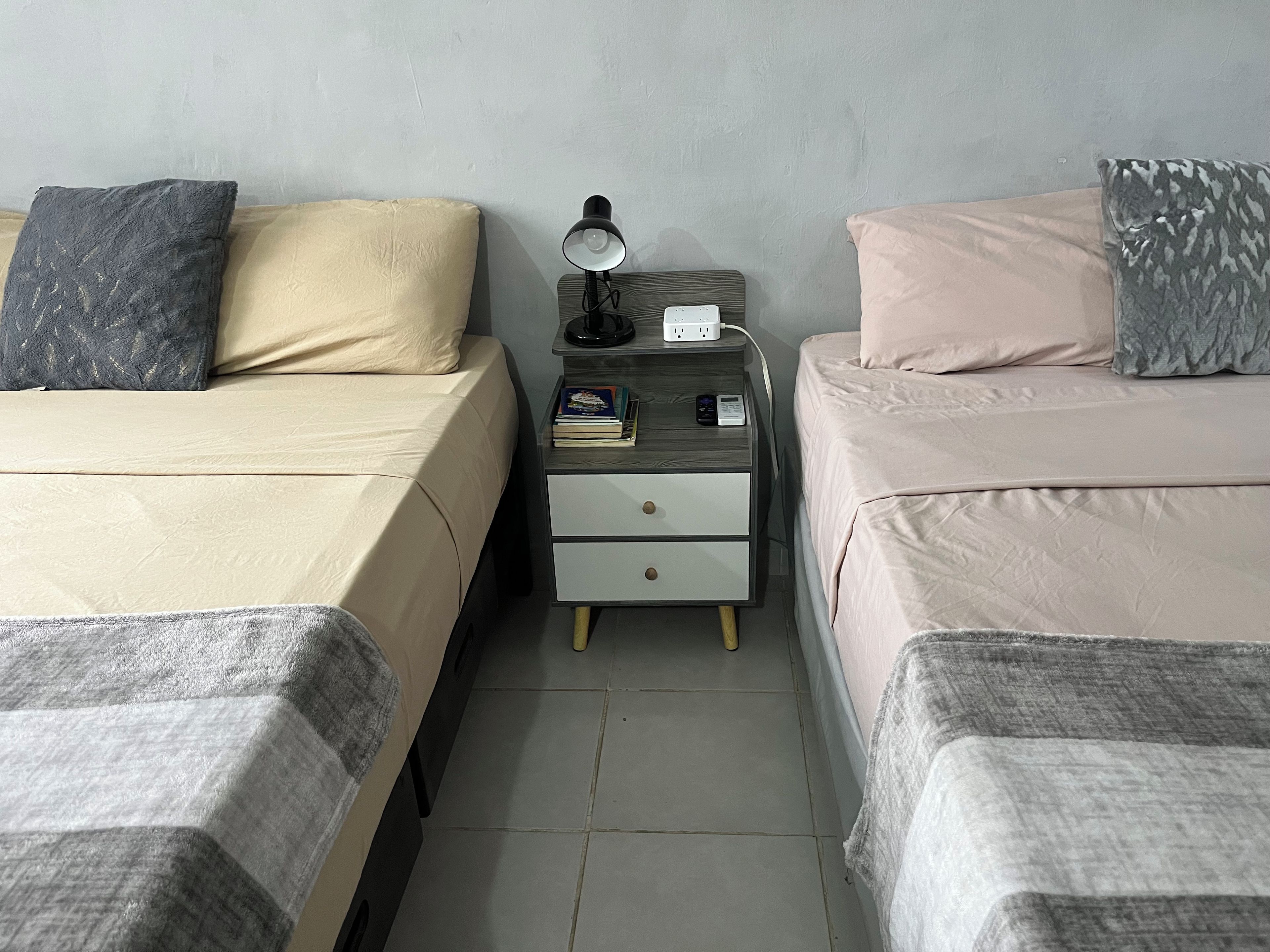Un dormitorio con dos camas, una mesita de noche con lámpara, libros y una regleta eléctrica.