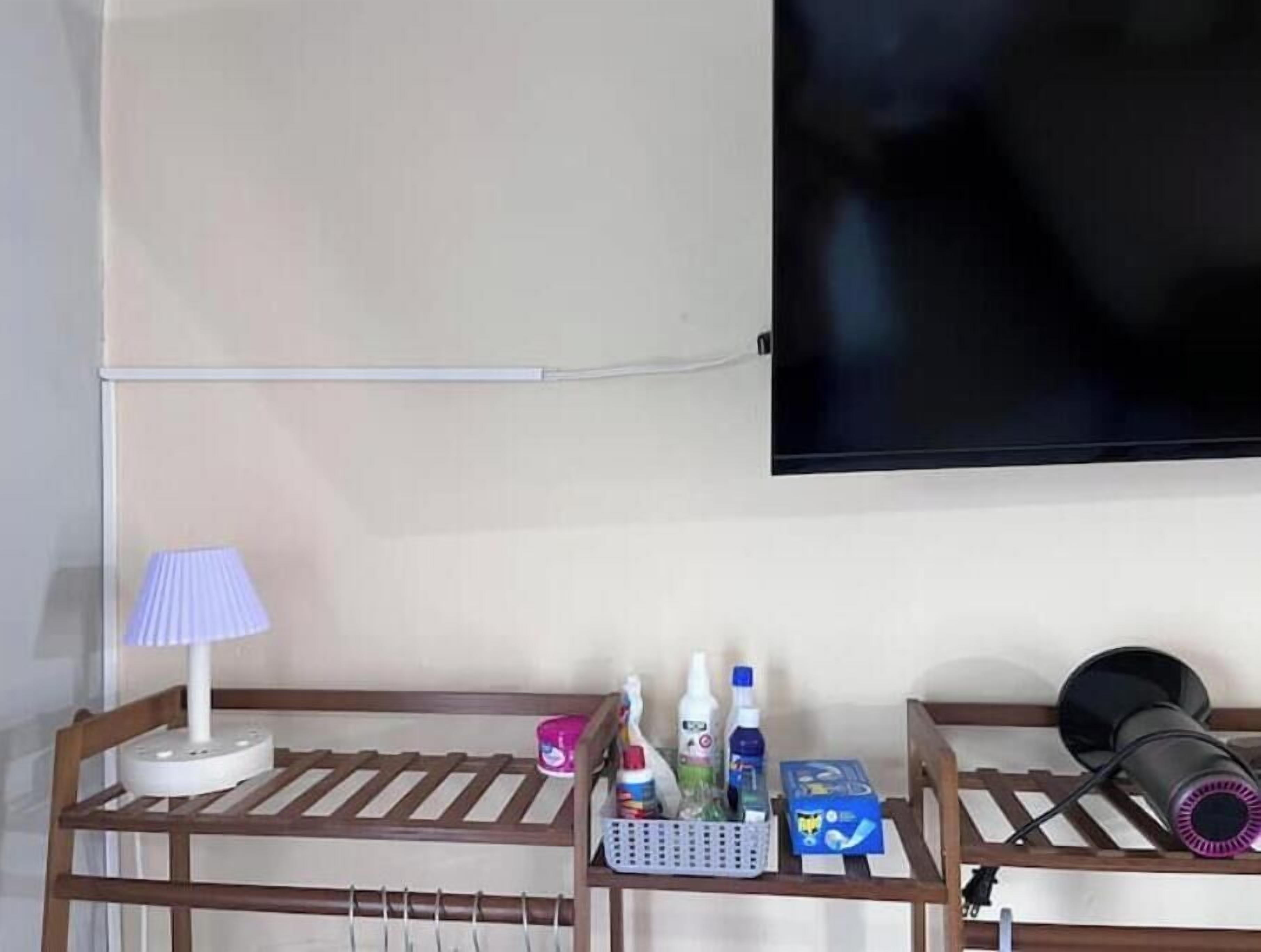 Habitación de hotel con televisión montada en la pared, mesita de noche de madera con lámpara, secador de pelo y artículos de aseo.