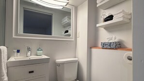 Een badkamer met toilet, wastafel, spiegel, planken met handdoeken en een prullenbak.