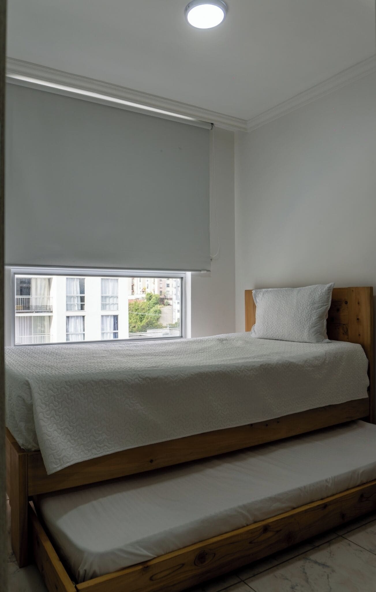 Ein Einzelbett mit Holzrahmen und einer Matratze, ein Fenster mit Blick auf Gebäude und eine Deckenleuchte.