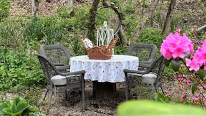 Ein runder Tisch mit einer blumigen Tischdecke, umgeben von Korbstühlen, in einem Garten voller Grün und Blumen.