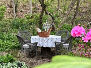 Ein runder Tisch mit einer blumigen Tischdecke, umgeben von Korbstühlen, in einem Garten voller Grün und Blumen.