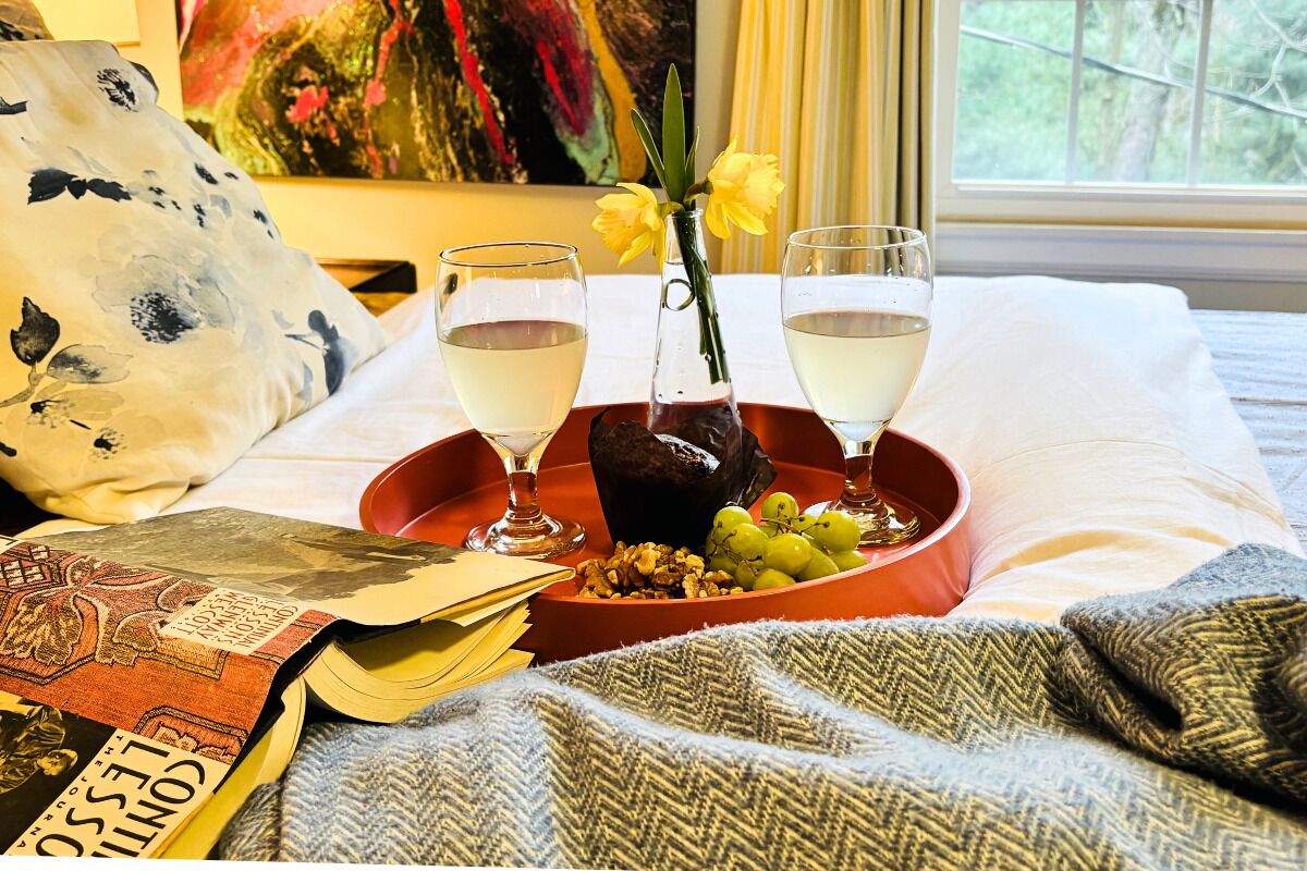 Un lit sur lequel se trouve un plateau rouge, avec deux verres de vin blanc, un vase de fleurs jaunes et un bol de raisins.