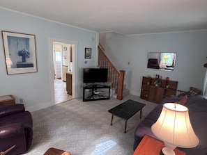 Una sala de estar con televisor, un sofá, una mesa de centro y una lámpara. Se ve una escalera que sube a otra habitación.