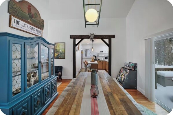 Un comedor con una larga mesa de madera, un mueble azul con puertas de vidrio y vista a la cocina por un arco abierto.